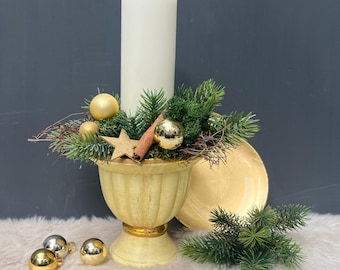 May include: A cream-colored pillar candle sits in a fluted yellow pedestal bowl, decorated with green pine branches, gold ornaments, a wooden star, and a cinnamon stick. Two additional gold ornaments and a pine sprig rest nearby.