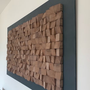 Puede incluir: Una pieza de arte de pared hecha de bloques de madera de varias formas y tama&ntilde;os. Los bloques est&aacute;n dispuestos en un patr&oacute;n geom&eacute;trico y est&aacute;n pintados en un color madera natural. La pieza de arte est&aacute; montada sobre un fondo gris oscuro.