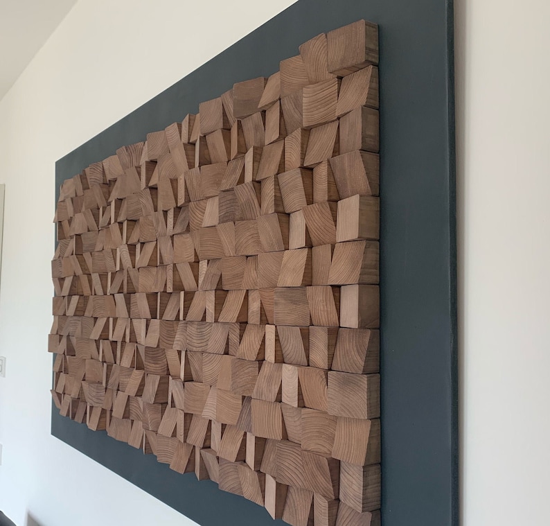 Puede incluir: Una pieza de arte de pared hecha de bloques de madera de varias formas y tama&ntilde;os. Los bloques est&aacute;n dispuestos en un patr&oacute;n geom&eacute;trico y est&aacute;n pintados en un color madera natural. La pieza de arte est&aacute; montada sobre un fondo gris oscuro.