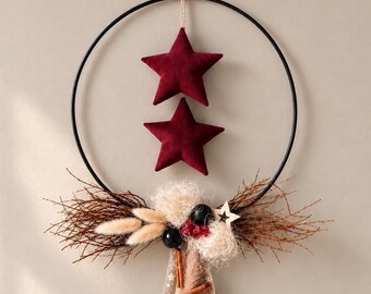 May include: A black metal hoop wall decoration featuring two hanging dark red fabric stars. The bottom of the hoop is adorned with brown twigs, beige dried grasses, small black spheres, red berries, and a beige ribbon with white stars.
