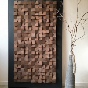 Puede incluir: Una gran pieza de arte de pared de madera hecha de muchos cuadrados peque&ntilde;os, dispuestos en un patr&oacute;n geom&eacute;trico. Los cuadrados son de color marr&oacute;n oscuro y tienen una veta de madera natural. El arte de pared est&aacute; enmarcado en un marco negro y est&aacute; colgado en una pared blanca. Un jarr&oacute;n con una rama seca est&aacute; en primer plano.