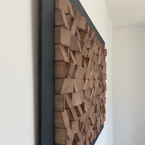 Puede incluir: Una escultura de pared hecha de bloques de madera de varios tama&ntilde;os y formas, dispuestos en un patr&oacute;n geom&eacute;trico. La escultura est&aacute; montada en un marco negro y tiene un acabado de madera natural.