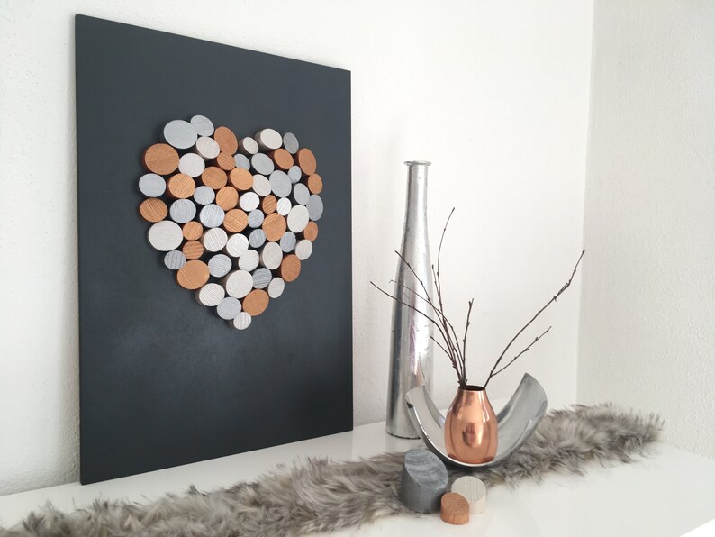 May include: A black canvas with a heart shape made of silver and copper colored circles. A silver vase with a branch of twigs is on a white surface with a gray fur rug. A copper vase with a branch of twigs is on a silver tray with two gray and copper colored circles.