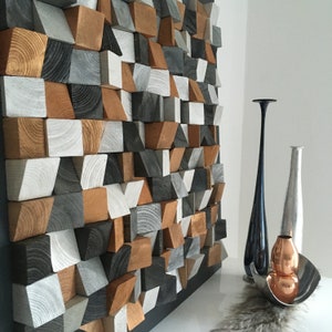 May include: A wall art piece made of geometric wooden blocks in shades of gray, white, and brown. The blocks are arranged in a random pattern, creating a modern and abstract design.