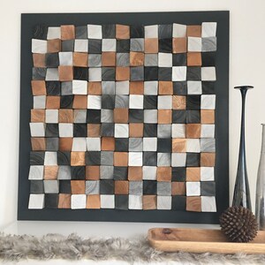 May include: A square wall art piece made of wood blocks in various shades of brown, gray, and white. The blocks are arranged in a checkered pattern and are slightly raised from the surface, creating a 3D effect. The art piece is framed in black and is hanging on a white wall.