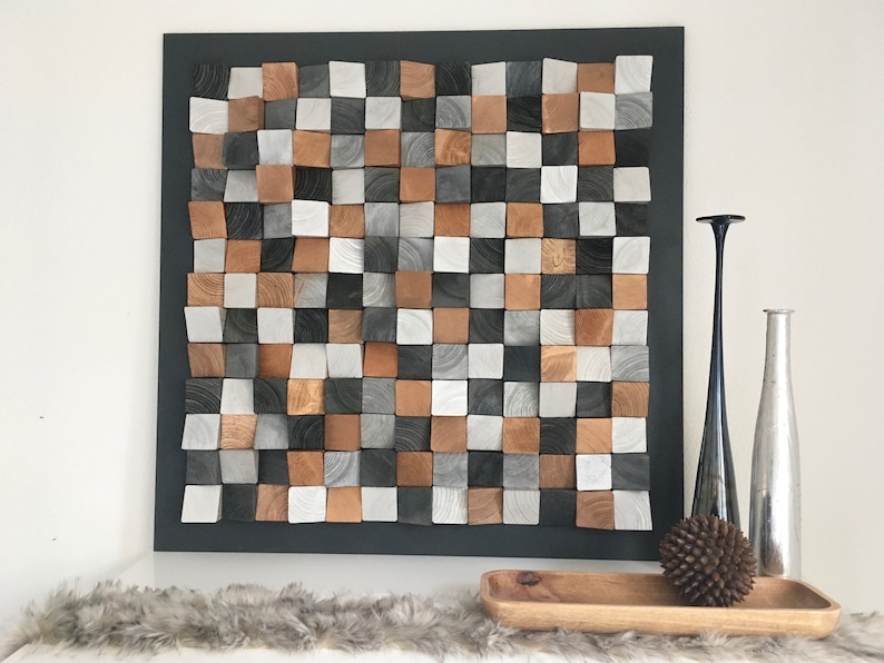 May include: A square wall art piece made of wood blocks in various shades of brown, gray, and white. The blocks are arranged in a checkered pattern and are slightly raised from the surface, creating a 3D effect. The art piece is framed in black and is hanging on a white wall.
