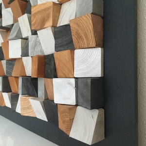 May include: A wall art piece made of wood blocks in various shades of gray, white, and brown. The blocks are arranged in a geometric pattern and are mounted on a black background.