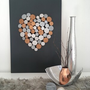 May include: A heart shape made of wooden circles painted in silver, gold, and copper tones is mounted on a dark gray background. A silver vase with a copper vase inside sits on a white surface with a gray fur rug.