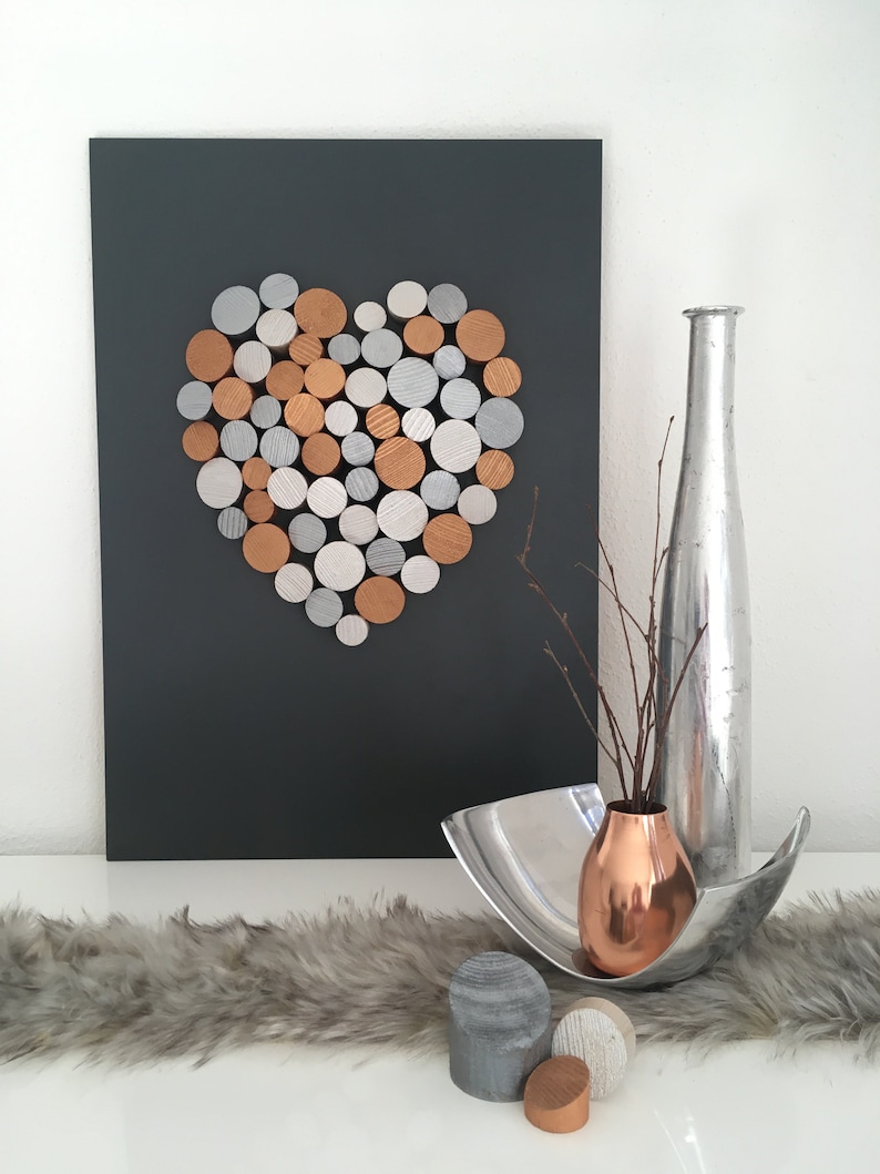 May include: A heart shape made of wooden circles painted in silver, gold, and copper tones is mounted on a dark gray background. A silver vase with a copper vase inside sits on a white surface with a gray fur rug.