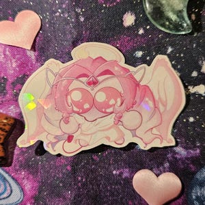May include: A pink sticker of a cartoon character with large eyes, wings, and a crown. The sticker has a holographic effect, surrounded by pink hearts and a space-themed background.