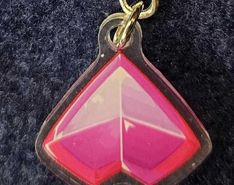 Könnte beinhalten: Ein klarer Acryl-Anhänger mit einem geometrischen Design in Rosa- und Weißtönen, umrandet in Schwarz. Der Anhänger ist an einem goldfarbenen Metallverschluss befestigt.