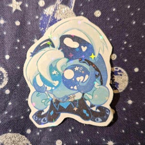 May include: A die-cut sticker depicting a cartoon character with blue and white hair and accents. The character has a large, round head and body with a whimsical design. The sticker has a holographic finish, with stars and other shapes. The background is a dark blue fabric.