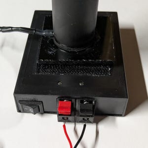 May include: A black electronic device with a cylindrical component and a rectangular base. The base has a power switch, red and black terminals, and a strip of Velcro. Red and black wires extend from the terminals.