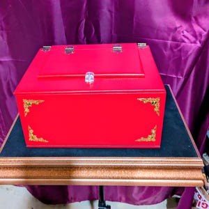 May include: A vibrant red wooden box with a hinged lid and a clear crystal clasp. The box features gold-coloured corner accents and rests on a black surface atop a decorative stand.
