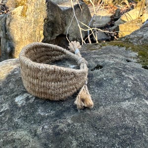 Op de afbeelding: Een geweven armband van natuurlijke vezels, waarschijnlijk jute of hennep, ligt op een getextureerde grijze rots. De armband is lichtbruin en heeft een eenvoudig, rustiek ontwerp. De uiteinden zijn samengebonden met een kwast.
