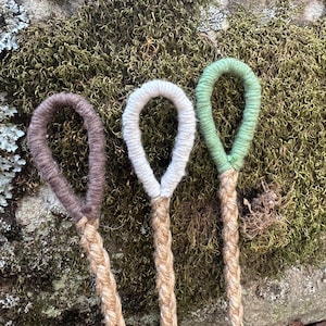 May include: Three handmade rope plant hangers with looped tops in brown, white, and green. The braided rope descends from the loops, showcasing a natural, rustic aesthetic. The background features moss and lichen on a stone surface.