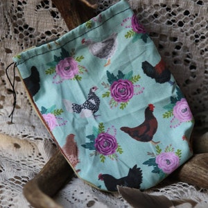 May include: A light blue fabric drawstring pouch featuring a repeating pattern of various chickens in shades of brown, black, and white, interspersed with pink and purple floral illustrations with green leaves and thin black drawstring cords.