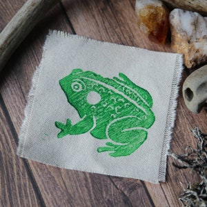 May include: A square piece of off-white fabric with frayed edges featuring a screen-printed illustration of a green frog in profile. The frog is depicted with textured skin, a circular eye, and a distinct circular marking behind its eye.