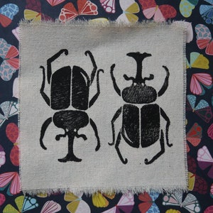 May include: A square piece of light beige, textured fabric with frayed edges featuring two black screen-printed beetle illustrations. The beetles are positioned side-by-side, each displaying distinct horn structures and segmented body shapes.