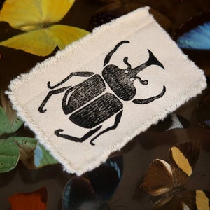 May include: A rectangular piece of off-white fabric with frayed edges featuring a black screen-printed illustration of a rhinoceros beetle. The fabric patch is positioned centrally, displaying a clear, detailed graphic of the insect's anatomy.