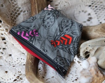 May include: A small zipped pouch featuring a grey fabric exterior with a repeating pattern of insects. The insects have black wings and bodies accented with bright pink and red details. The pouch has a black zip and a solid red interior lining.