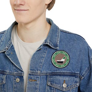 Op de afbeelding: Een blauwe denimjas met een ronde patch met de omtrek van de Verenigde Arabische Emiraten in groen, wit en zwart, omringd door de tekst "UAE * UAE * UAE * UAE * UAE * UAE" in zwart.