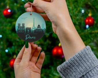 May include: A round, white Christmas bauble featuring a blue and green illustration of the Paris skyline, including the Eiffel Tower, Notre Dame Cathedral, and the Arc de Triomphe. The text "Welcome to France" and "Paris" is printed on the bauble.