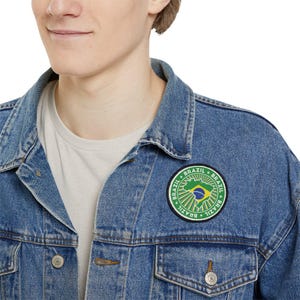 Puede incluir: Una chaqueta vaquera azul con un parche circular verde, amarillo y azul que dice "Brasil" cuatro veces alrededor del borde. El parche presenta un contorno estilizado de Brasil con la bandera brasileña en su interior.