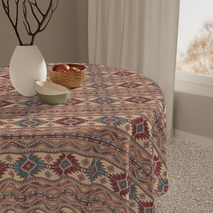 May include: A round tablecloth with a Southwestern-inspired pattern in shades of beige, red, and blue. The intricate design features geometric shapes and tribal motifs, creating a warm and inviting aesthetic. A white vase, a small basket with fruit, and two bowls are on the table.