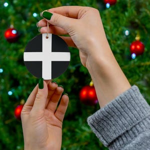 May include: A round Christmas ornament with a black and white design. The ornament features a white cross on a black background. The ornament is being held by a hand in front of a Christmas tree.