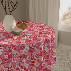 Op de afbeelding: Een ronde tafelkleed met een patchwork-ontwerp met harten, enveloppen en andere Valentijnsdag-thema's in roze en rode tinten. Een witte vaas, een rieten mand met fruit en twee kommen staan op de tafel.