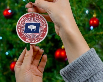 May include: A red, white, and blue Christmas ornament with the word "WYOMING" written around the edge and a silhouette of a bison in the center. The ornament is being held by a hand with green nail polish.