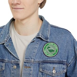 Puede incluir: Una chaqueta vaquera azul con un parche circular verde y blanco que dice "Arabia Saudita" con un mapa de Arabia Saudita dentro del círculo.