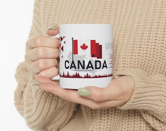 May include: White ceramic mug featuring a red and white Canadian flag design and the word "CANADA" in black text. The mug is being held by a person wearing a beige sweater.
