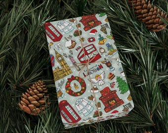 May include: A wrapped gift with a light blue background and a repeating pattern of London-themed illustrations, including a red telephone booth, Big Ben, and a double-decker bus. The gift is tied with twine and set on a bed of pine needles and pine cones.