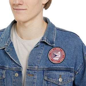 May include: A light blue denim jacket with a round Ohio state flag patch. The patch features a red border with the word "OHIO" and a white and red design with a blue triangle.