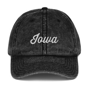 Könnte beinhalten: Eine schwarze Baseballkappe im Vintage-Stil mit dem Wort "Iowa" in weißer Schrift auf der Vorderseite gestickt. Die Kappe hat einen gebogenen Schirm und einen Knopf oben. Der Stoff hat ein verblasstes, abgenutztes Aussehen.