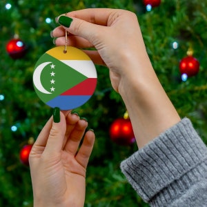 May include: A hand holds a round Christmas ornament with the flag of Comoros. The ornament is yellow, green, red, and blue. The flag has a crescent moon and four stars.
