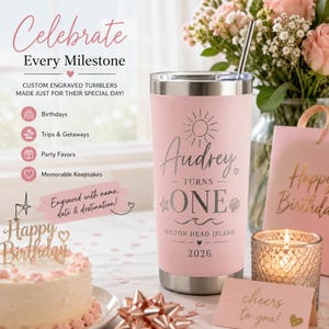 May include: A pink custom engraved tumbler with a silver lid and straw. The tumbler is personalized with the name "Audrey", the words "turns one", and the location "Hilton Head Island 2026". The image also includes a cake, gift bag, and candle.