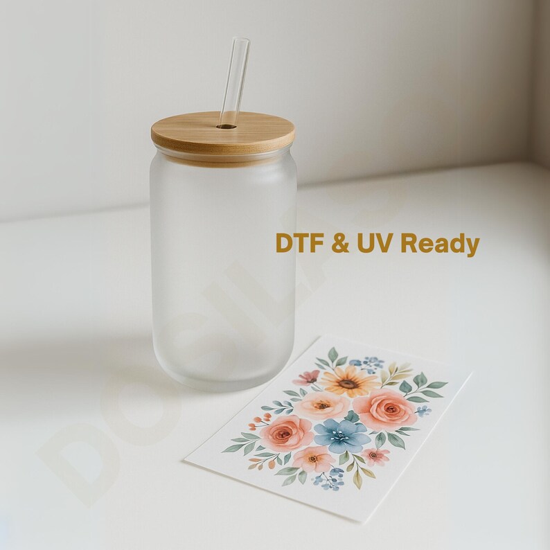 May include: A frosted glass can-shaped tumbler with a bamboo lid and a clear glass straw. A floral design card with peach, blue, and yellow flowers is next to the tumbler. The text "DTF & UV Ready" is printed on the glass.