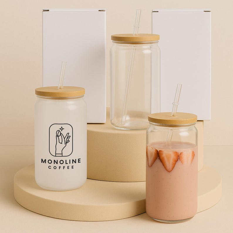 May include: Three glass tumblers with bamboo lids and clear straws. One tumbler is frosted with a black and white "MONOLINE COFFEE" logo. Another contains a pink smoothie with strawberry slices. White boxes are in the background.