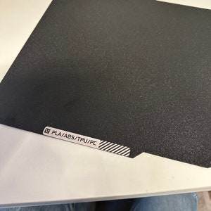 K&ouml;nnte beinhalten: Ein schwarzes, strukturiertes Druckbett f&uuml;r einen 3D-Drucker. Das Bett tr&auml;gt ein Etikett mit der Aufschrift "PLA/ABS/TPU/PC".