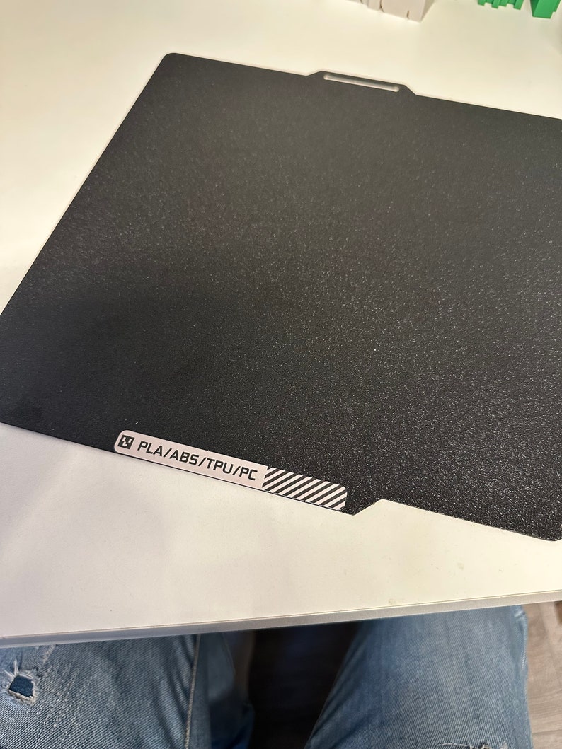 K&ouml;nnte beinhalten: Ein schwarzes, strukturiertes Druckbett f&uuml;r einen 3D-Drucker. Das Bett tr&auml;gt ein Etikett mit der Aufschrift "PLA/ABS/TPU/PC".