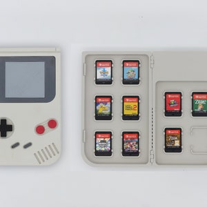 Può includere: Una console per videogiochi portatile Game Boy Color bianca accanto a una custodia per giochi Nintendo Switch bianca con 10 cartucce di gioco all'interno. Le cartucce sono per giochi come Pokemon, Mario Party e Zelda.