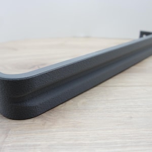 May include: A dark gray, textured metal bumper section with a curved corner. The bumper has a rectangular shape with a slightly raised central section. The object is set against a light wood grain surface.