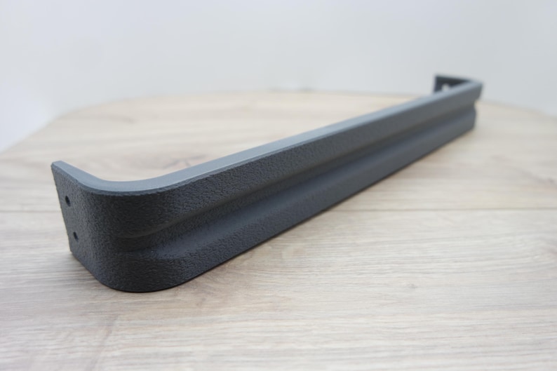 May include: A dark gray, textured metal bumper section with a curved corner. The bumper has a rectangular shape with a slightly raised central section. The object is set against a light wood grain surface.