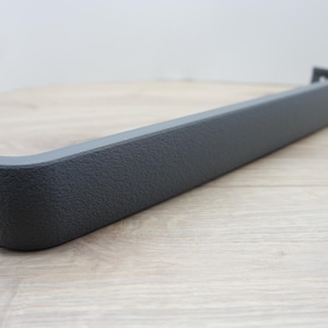 May include: A dark gray, textured plastic automotive part. The piece is long and rectangular with a curved end and a small, angled extension. The part appears to be new and unused, with a matte finish.