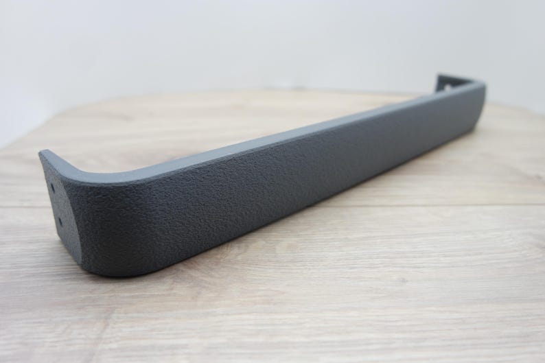 May include: A dark gray, textured plastic automotive part. The piece is long and rectangular with a curved end and a small, angled extension. The part appears to be new and unused, with a matte finish.