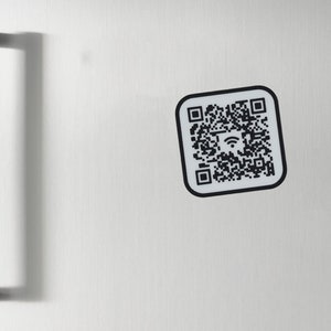 Könnte beinhalten: Ein schwarz-weißer QR-Code-Aufkleber auf einer Edelstahl-Kühlschranktür. Der QR-Code hat ein WLAN-Symbol in der Mitte.