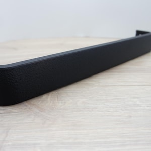 May include: A black, textured, rectangular automotive part with a curved end and two holes. The part is set against a light wood-grain surface and a plain white background. The part appears to be made of a durable material.